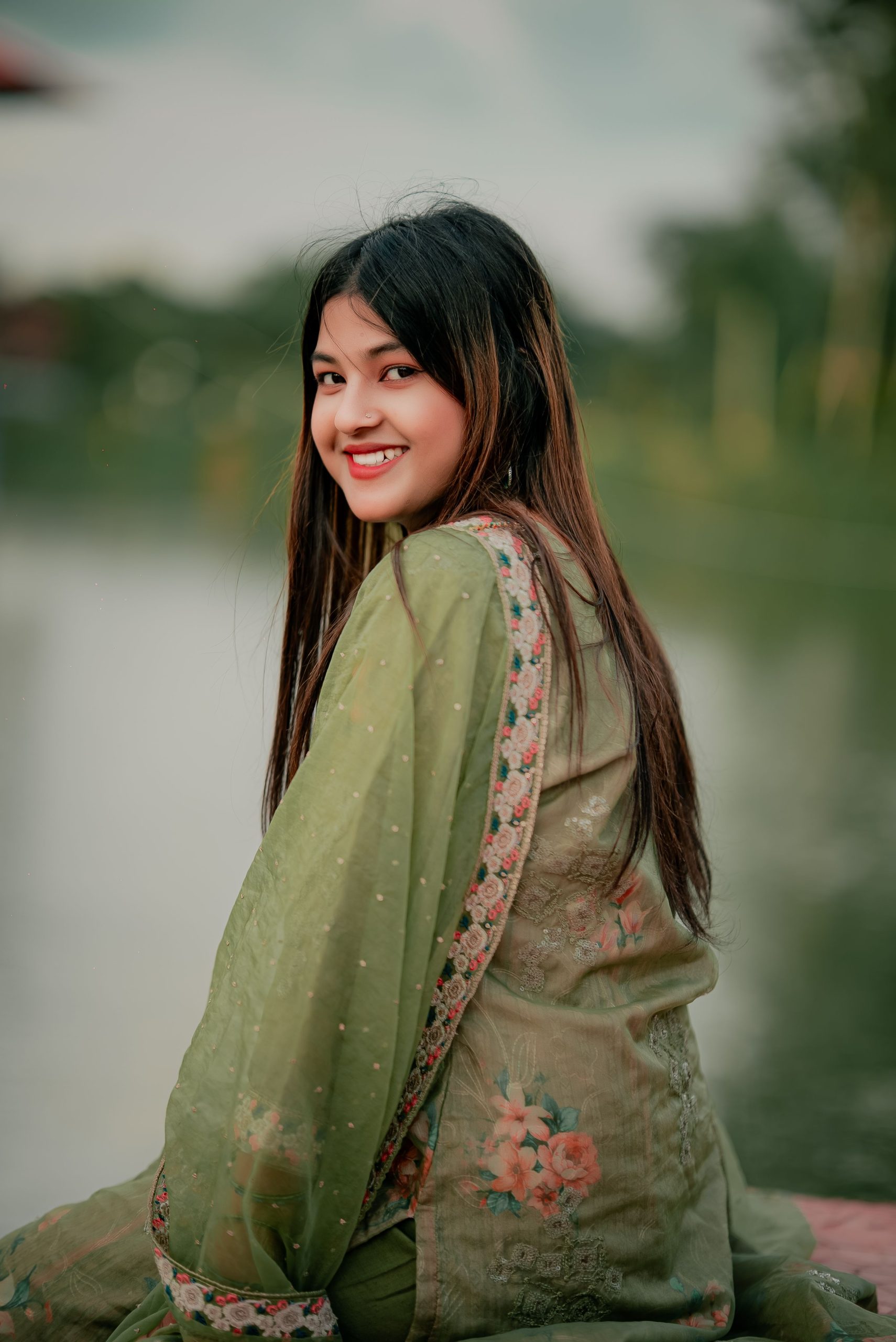 A smiling woman in traditional green attire with floral embroidery by a serene lake in Bangladesh.